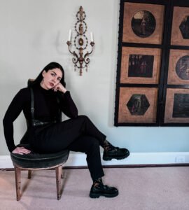 Woman sits in a chair wearing an all black gothic outfit paired with chunky loafers and leather harness.